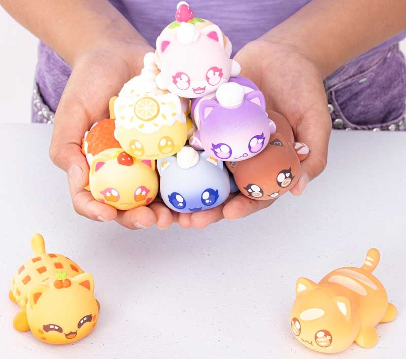Hands holding Aphmau MeeMeow Bakery squishy cat figures, colourful collectible sensory toys