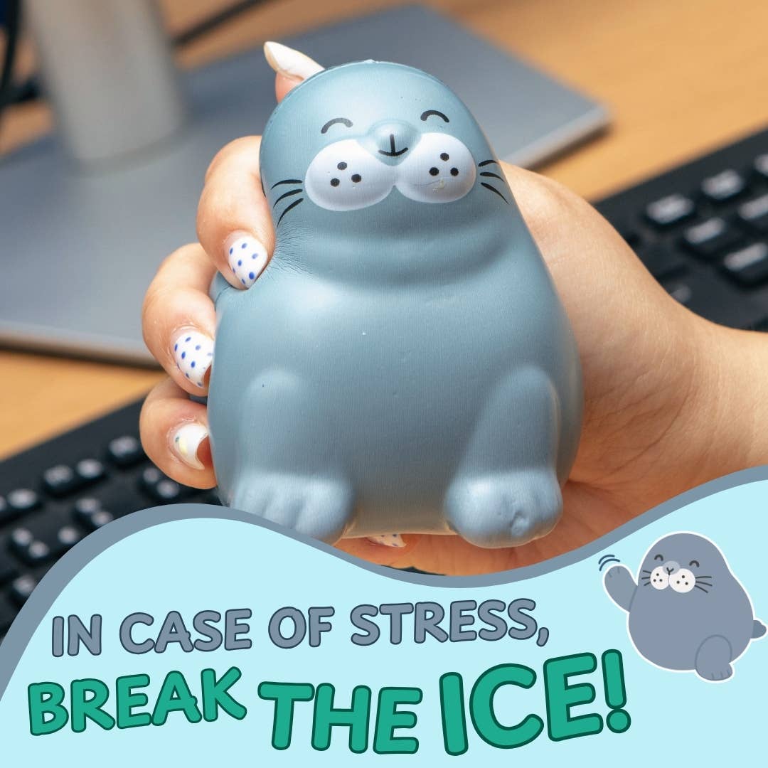 A hand holds a gray seal-shaped stress toy with a cartoon face. The background shows a desk with a keyboard and monitor, and a graphic overlay at the bottom reads 'IN CASE OF STRESS, BREAK THE ICE!' with a small seal illustration.