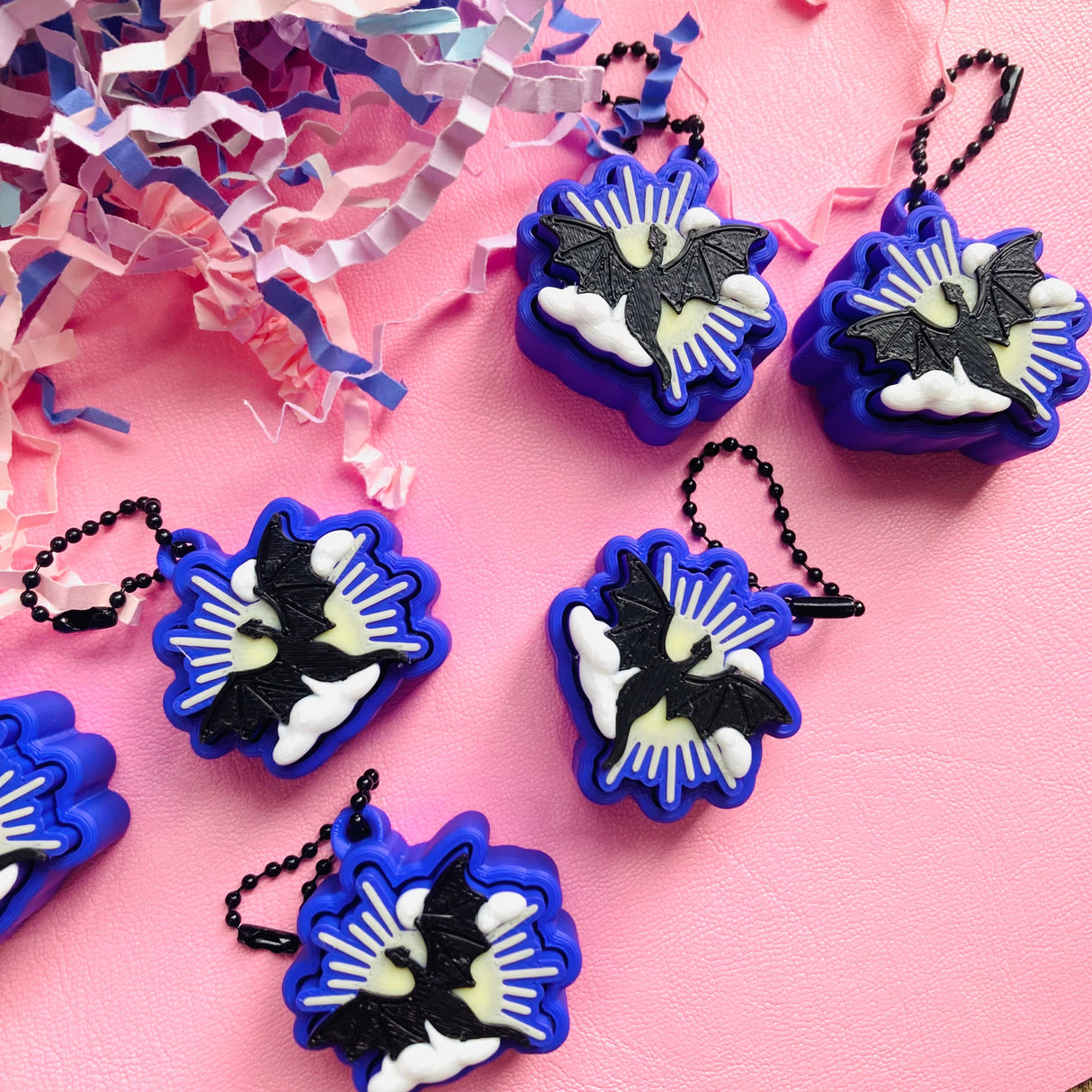 Multiple blue and black dragon clicker fidget keychains laid out on a pink surface with confetti