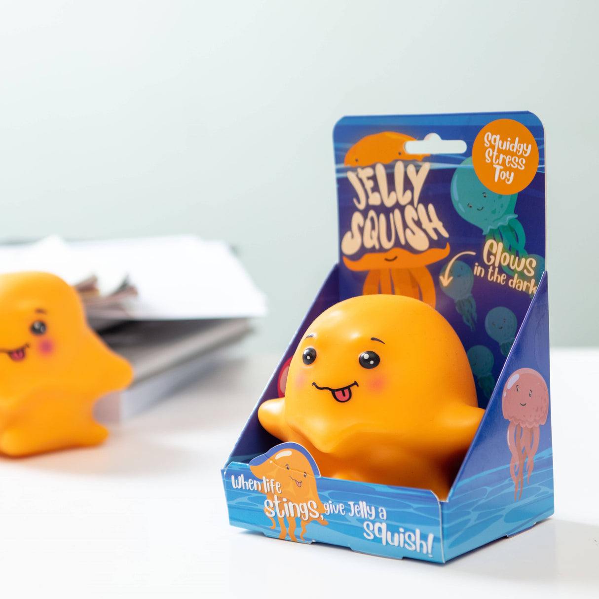 Orange kawaii jellyfish squishy stress toy next to its purple Jelly Squish gift box on a white desk