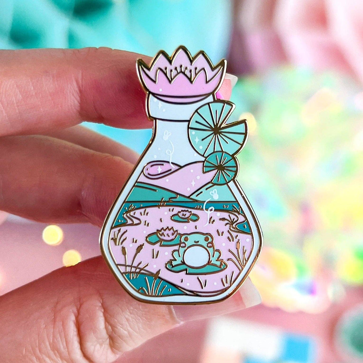 Lula Rocks Bottled Frog UV Glow enamel pin, pastel pink teal, hand-held close-up
