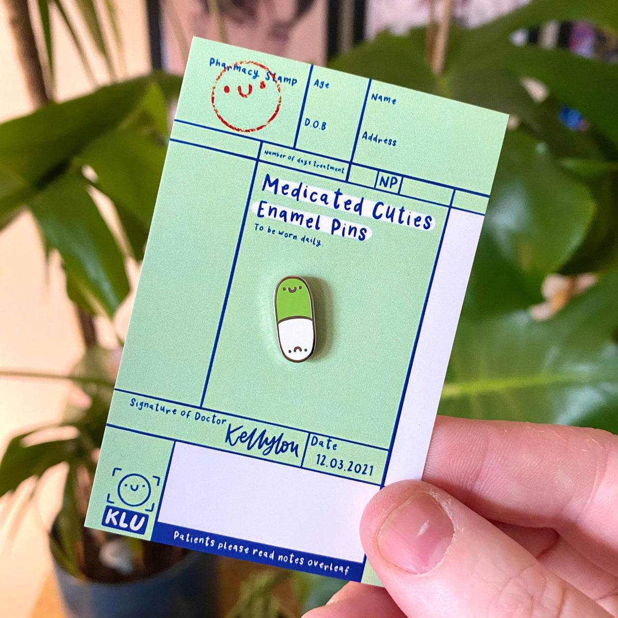 Green enamel pin on a card labeled 'Medicated Cuties Enamel Pins' held by a hand with a blurred green plant background.