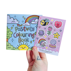Adult hand holding The Positivity Colouring Book and Positivity Sticker Sheet together on a white background
