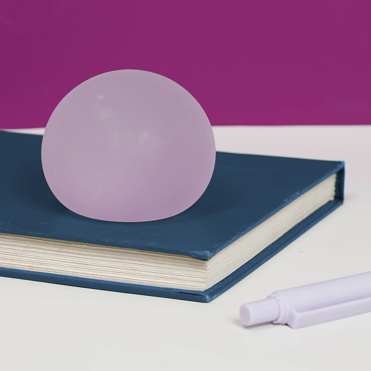Lilac Mist Speks Gump Original stress ball on a dark blue book beside a pen against a purple background