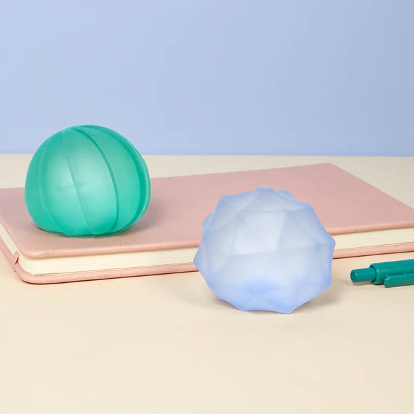 Teal and pale blue Speks Gump Surface stress balls displayed on a notebook beside pens on a desk