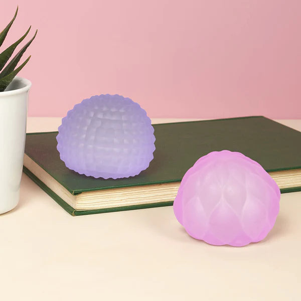 Purple and pink Speks Gump Surface stress balls displayed on a book beside a plant on a desk