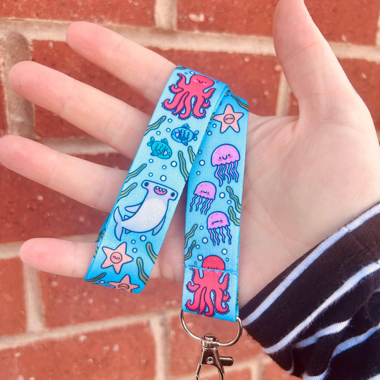 Hand holding a blue lanyard with pink and white sea creature designs against a brick wall.