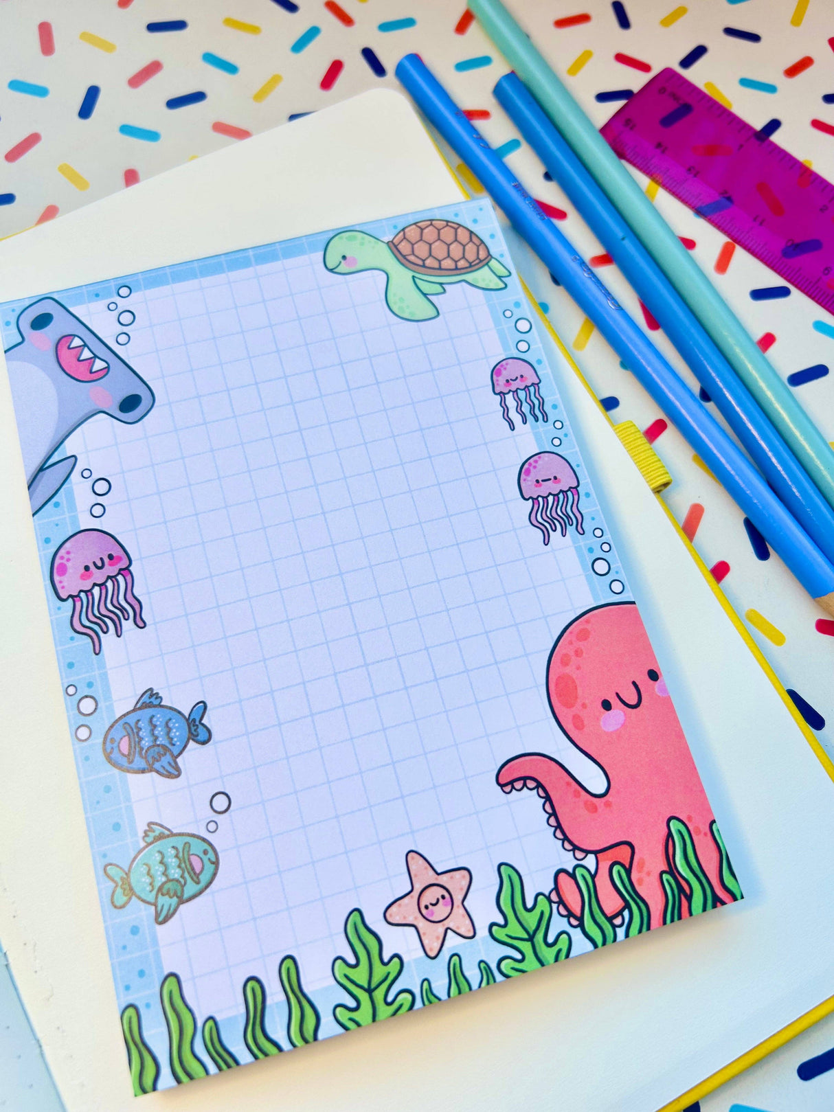 Under the Sea kawaii notepad laid flat on a desk beside blue pencils and a pink ruler showing the grid paper and ocean character border