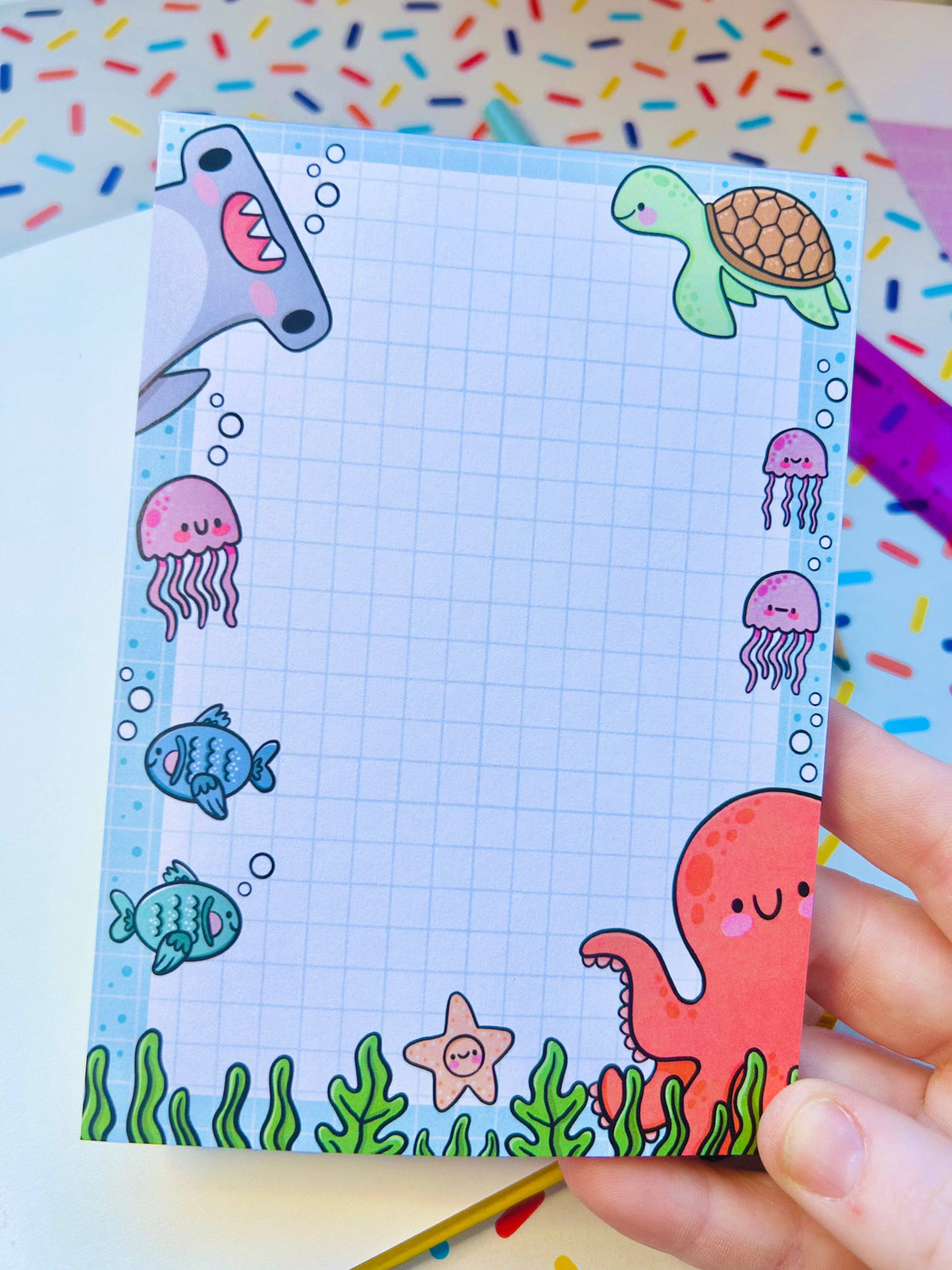 Adult hand holding the Under the Sea kawaii notepad against a colourful sprinkle-print background showing full illustration border
