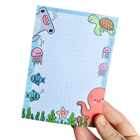 Adult hand holding the Under the Sea kawaii notepad on a white background showing the full octopus, shark, jellyfish and turtle border