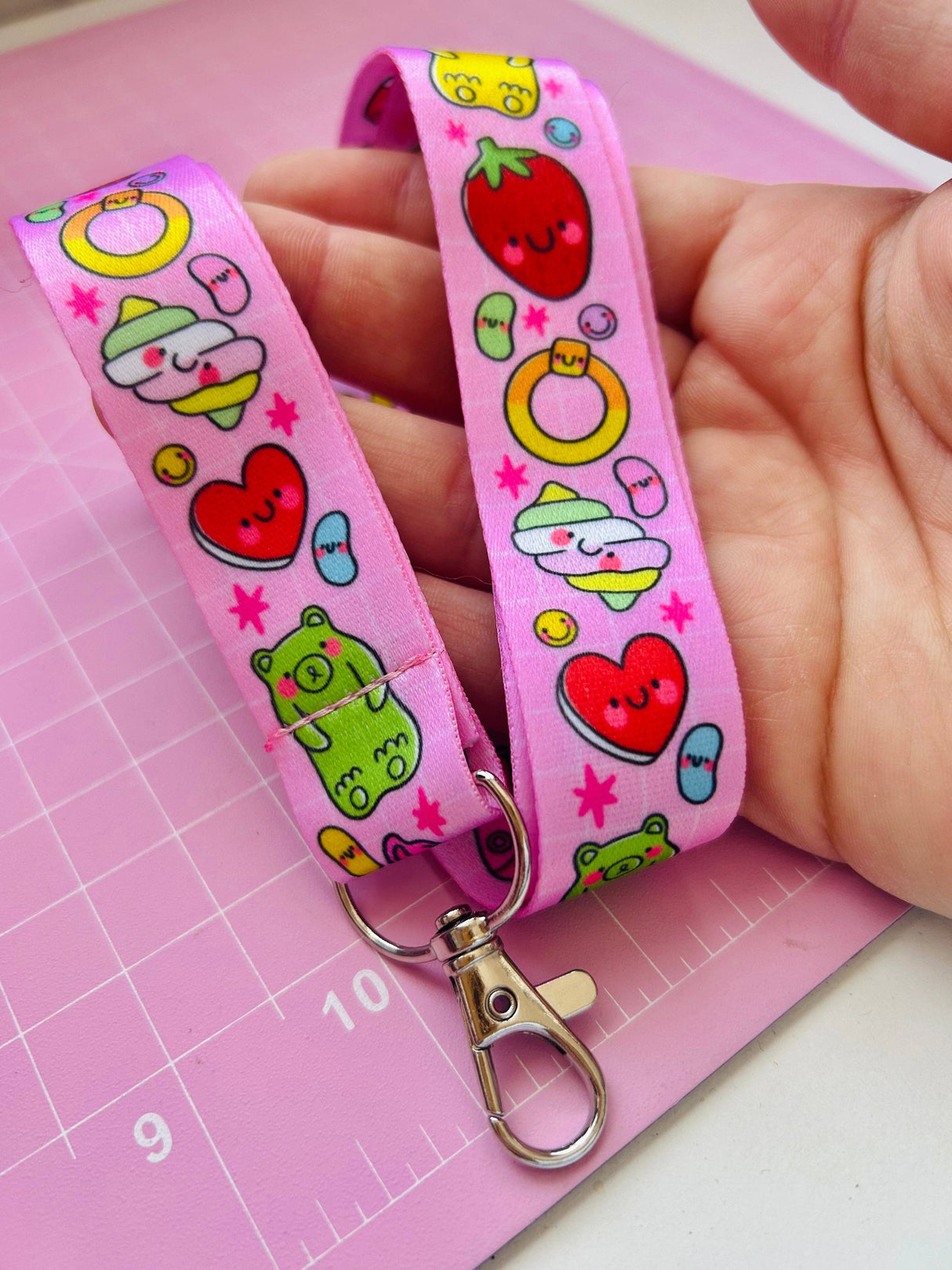 Adult hand holding the Pick 'n' Mix kawaii lanyard over a pink cutting mat showing the full print with gummy bears, strawberries and hearts