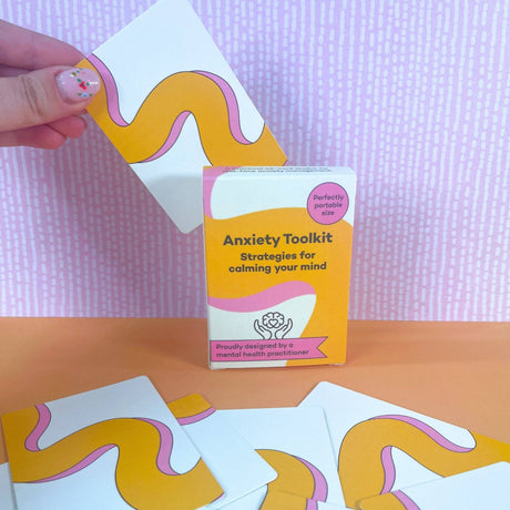 Hand with painted nails pulling a card from the Anxiety Toolkit deck, with more cards spread on an orange surface