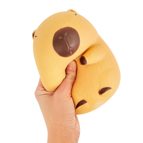The Big Squij Capybara slow-rise squishy toy being squeezed and deformed in one hand, showing the soft texture and large size of the yellow-orange capybara squishy against a white background
