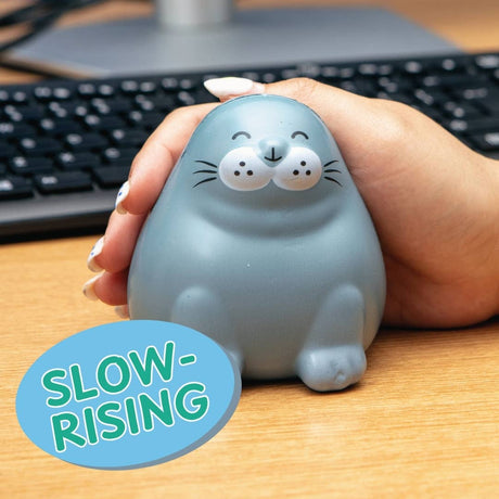 A person's hand holding a light blue seal-shaped stress toy on a wooden desk with a keyboard in the background. A blue circular sticker overlay in the bottom left reads 'SLOW-RISING'.