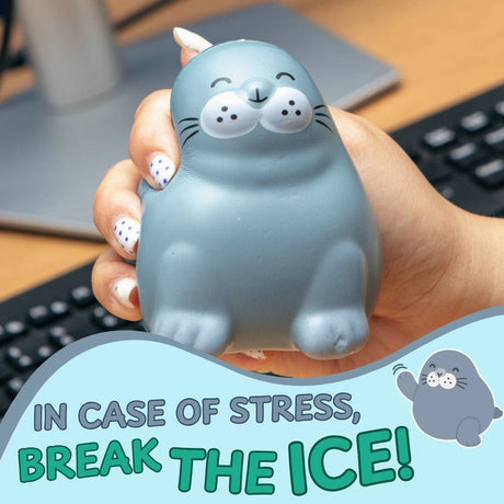 A hand holds a gray seal-shaped stress toy with a cartoon face. The background shows a desk with a keyboard and monitor, and a graphic overlay at the bottom reads 'IN CASE OF STRESS, BREAK THE ICE!' with a small seal illustration.