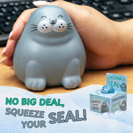 A person's hand holds a gray seal-shaped stress toy on a wooden desk with a keyboard in the background. The toy has a smiling face and small paws.