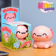 A pink axolotl-shaped squishy toy shown next to its blue illustrated retail box on a wooden surface, with a 'Squish Squad' logo in the top right.