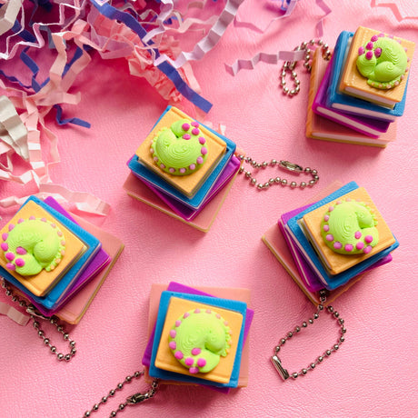 Multiple 3D printed sleeping dragon book stack clicker keychains on a pink surface with confetti