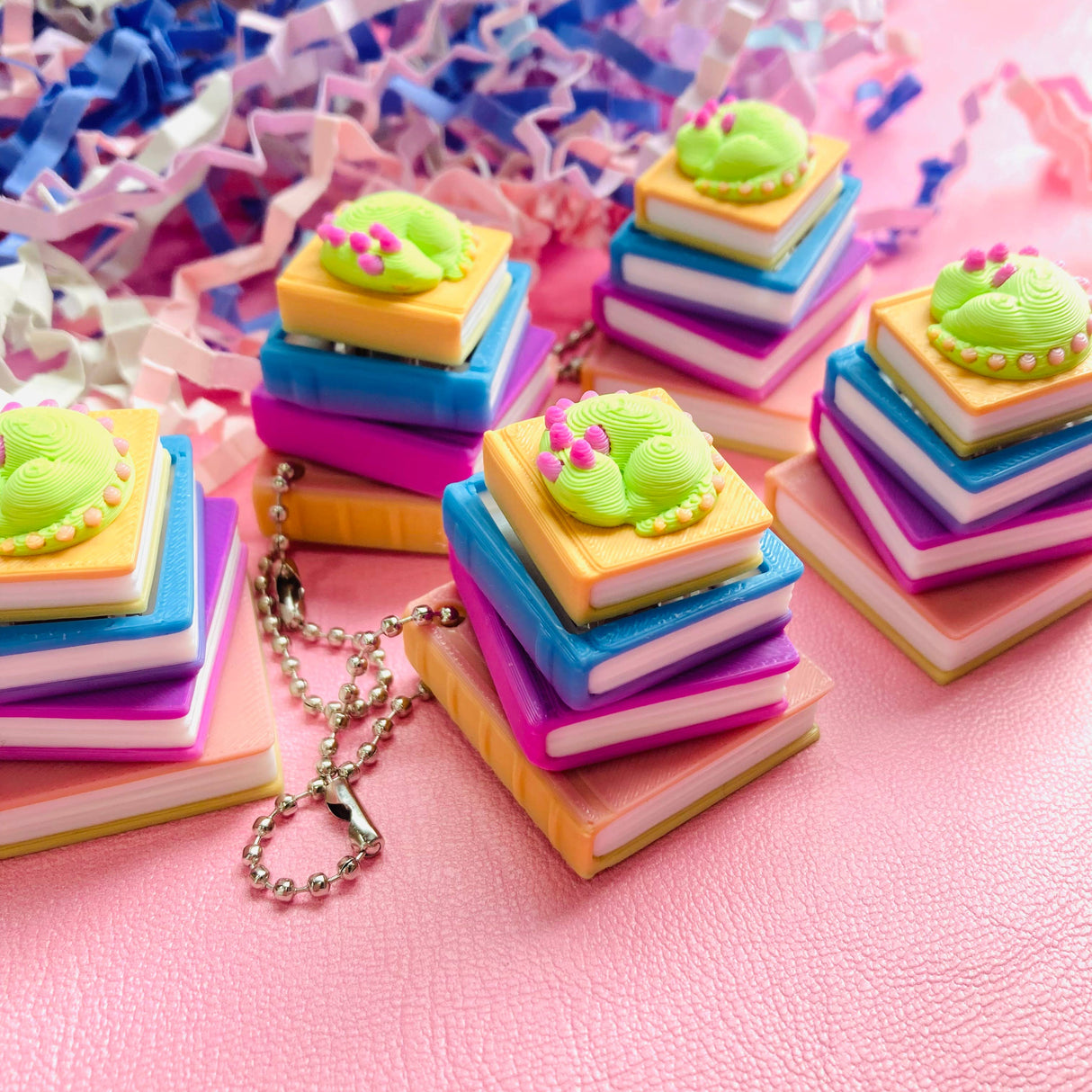 Five sleeping dragon book stack clicker keychains spread across a pink surface with confetti