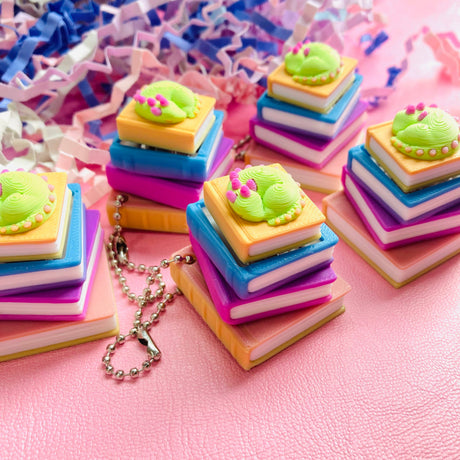 Five sleeping dragon book stack clicker keychains spread across a pink surface with confetti
