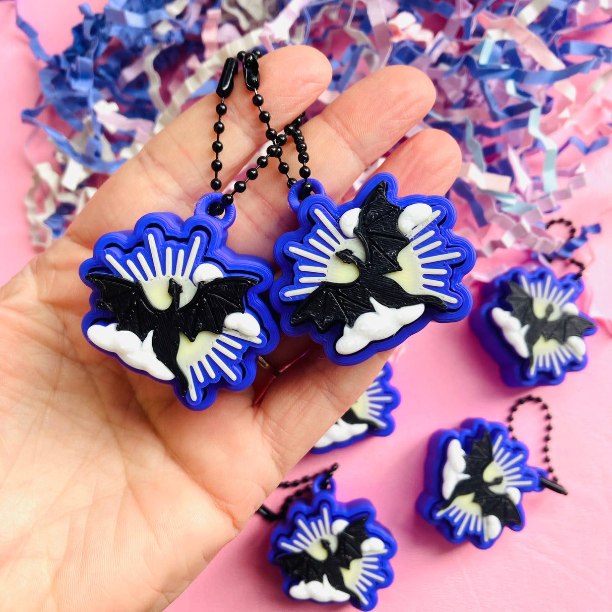 Two blue and black 3D printed dragon clicker fidget keychains held in a palm with pink confetti background