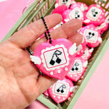 Single pink heart clicker keychain held in a palm in front of basket showing size and cherry pixel art