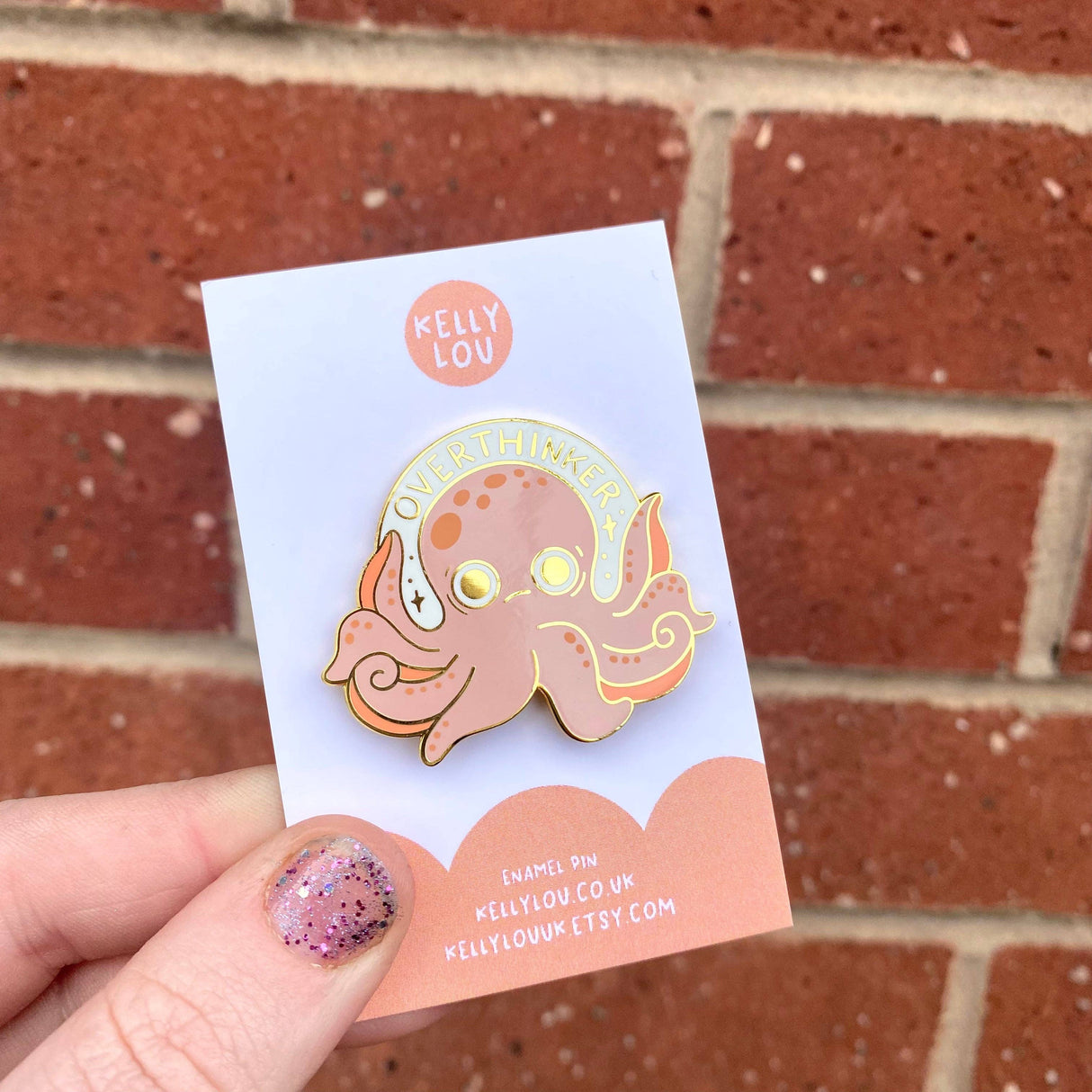 Pink octopus enamel pin on a Kelly Lou branded card against a brick wall.