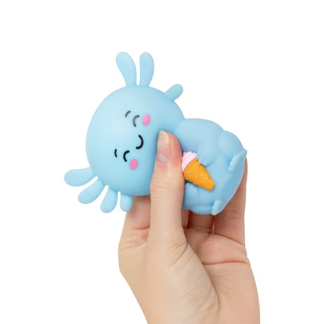 A light blue squishy toy character with a cute face and small antennae, being held by a person's hand. The character is holding a small orange and pink ice cream cone.