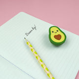 Legami avocado shaped eraser with heart pit resting on an open notebook beside a pencil
