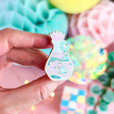 Lula Rocks Bottled Frog UV Glow enamel pin, pastel pink teal, hand angled view
