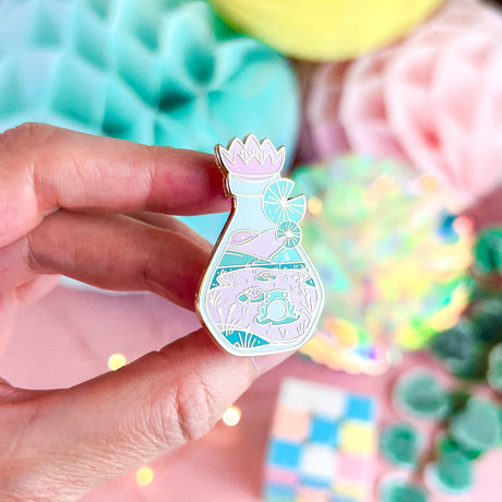 Lula Rocks Bottled Frog UV Glow enamel pin, pastel pink teal, hand angled view