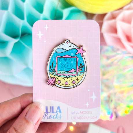 Colorful enamel pin on a pink card with 'Lula Rocks' branding, held against a vibrant background.