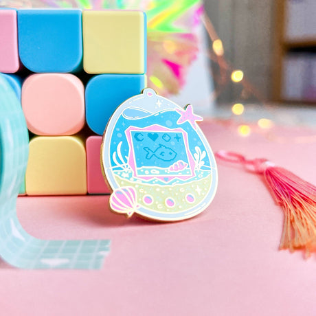 Pastel-colored enamel pin with a decorative charm featuring a fish design on a pink surface.