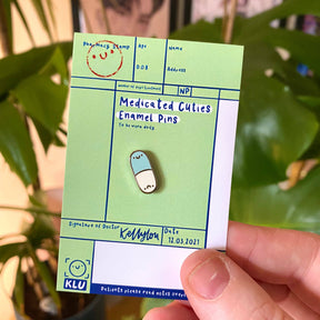 Medicated cuties enamel pin on a green card with a plant in the background