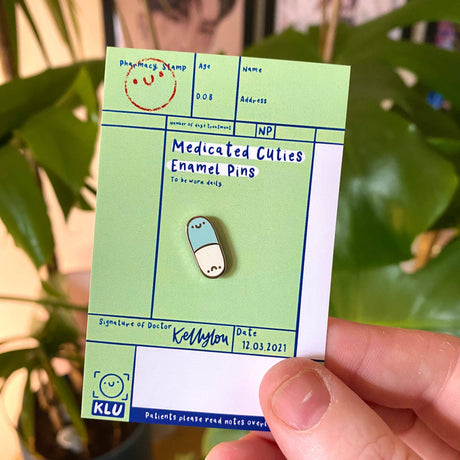 Medicated cuties enamel pin on a green card with a plant in the background