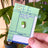 Green enamel pin on a card labeled 'Medicated Cuties Enamel Pins' held by a hand with a blurred green plant background.