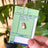 Medicated Cuties Enamel Pins packaging held by a hand with green leaves in the background