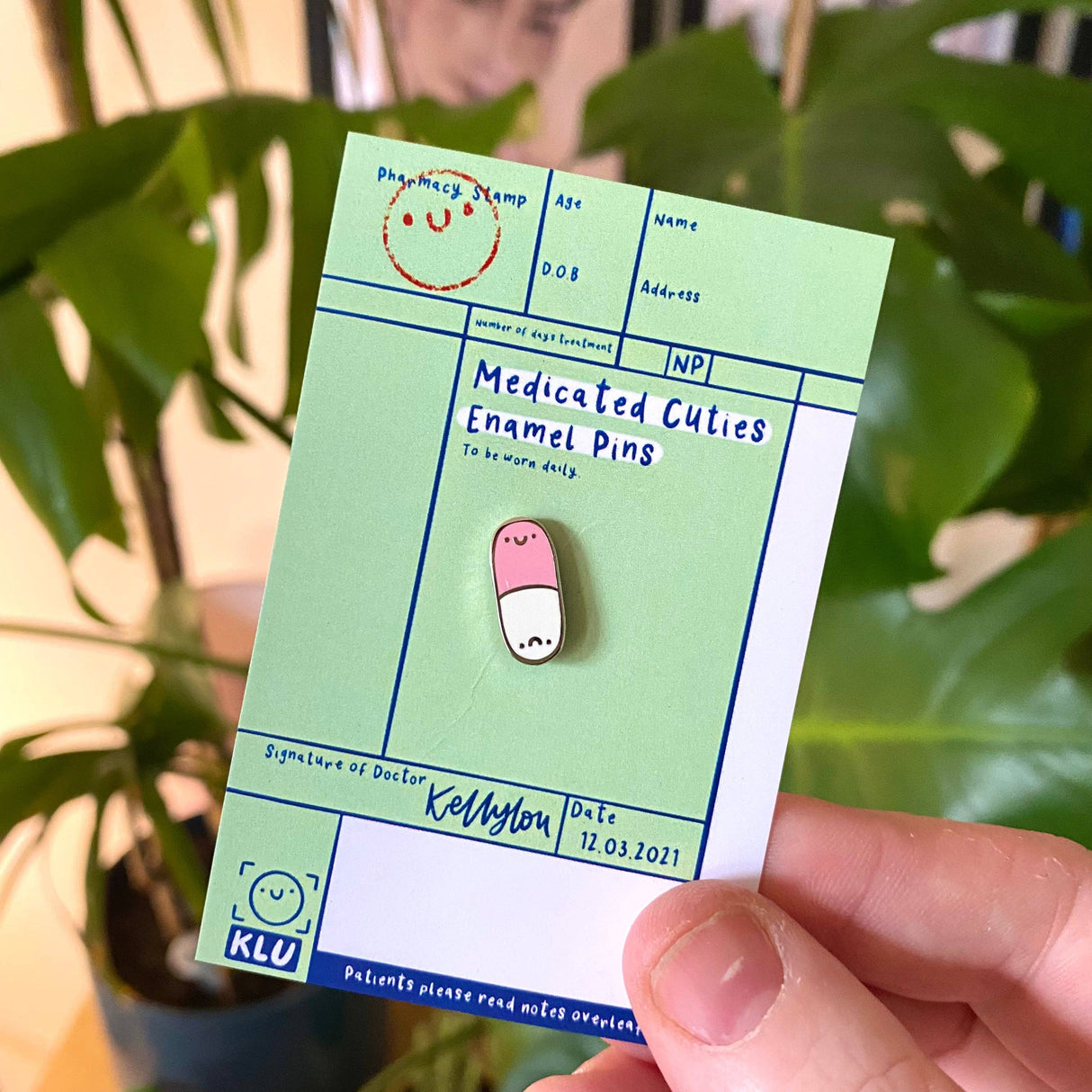 Medicated Cuties Enamel Pins packaging held by a hand with green leaves in the background
