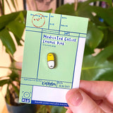 Medicated Cuties Enamel Pins on a green card held by a hand with a blurred green leafy background
