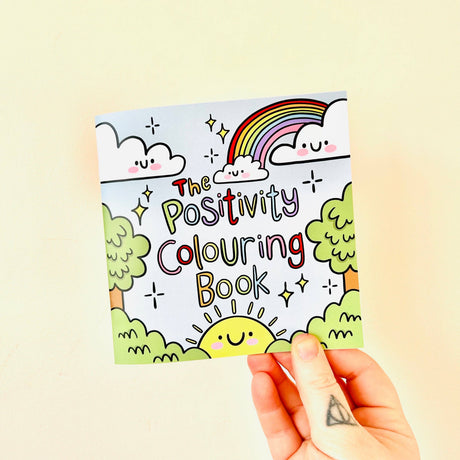 Adult hand holding The Positivity Colouring Book showing the full kawaii rainbow sun and cloud cover illustration