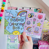 Adult hand holding The Positivity Colouring Book alongside the lilac Positivity Sticker Sheet in a colourful room setting
