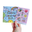 Adult hand holding The Positivity Colouring Book and Positivity Sticker Sheet together on a white background