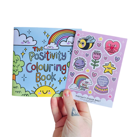 Adult hand holding The Positivity Colouring Book and Positivity Sticker Sheet together on a white background