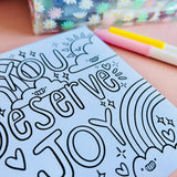 Uncoloured Positivity Colouring Book page reading You Deserve Joy with rainbow and sun illustration, shown beside pastel pens