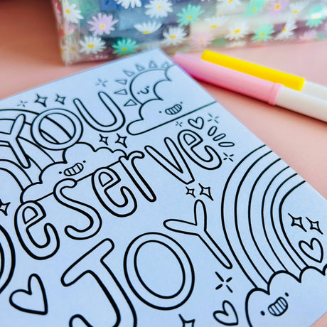 Uncoloured Positivity Colouring Book page reading You Deserve Joy with rainbow and sun illustration, shown beside pastel pens