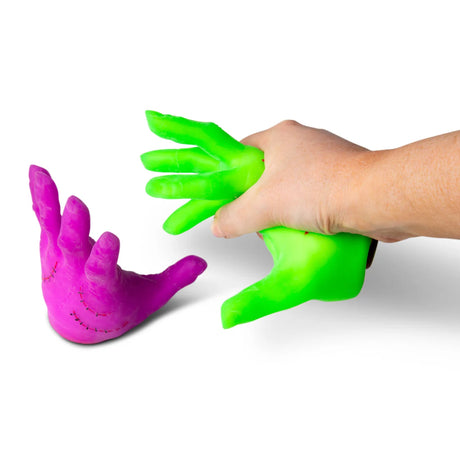 Adult hand squeezing a green Monster Hand alongside a pink one, showing the stretchy sand-filled texture