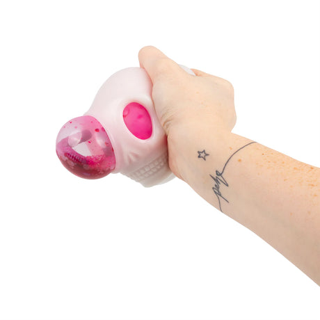 Adult hand squeezing a pale pink glow-in-the-dark skull squishy, with the pink dome bulging from the eye socket