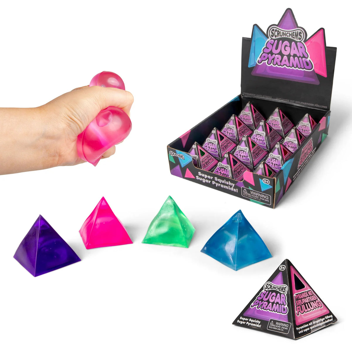 Hand squeezing a pink Scrunchems Sugar Pyramid beside its display box and four loose colours