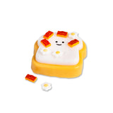 Smiling Scrunchems toast squishy covered in sticky bacon and fried egg pieces with loose accessories beside it