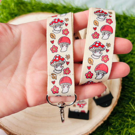 Adult hand holding the Shroom Cuties lanyard draped over a wooden slice showing mushroom and flower print detail with swivel clip